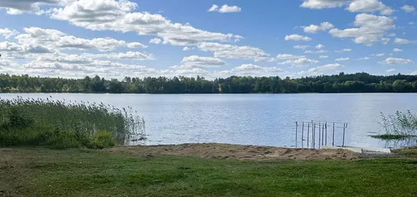 Sandstrand och badbrygga vid Lyttersta badplats. Sjö, vass och trädridå syns i bakgrunden.